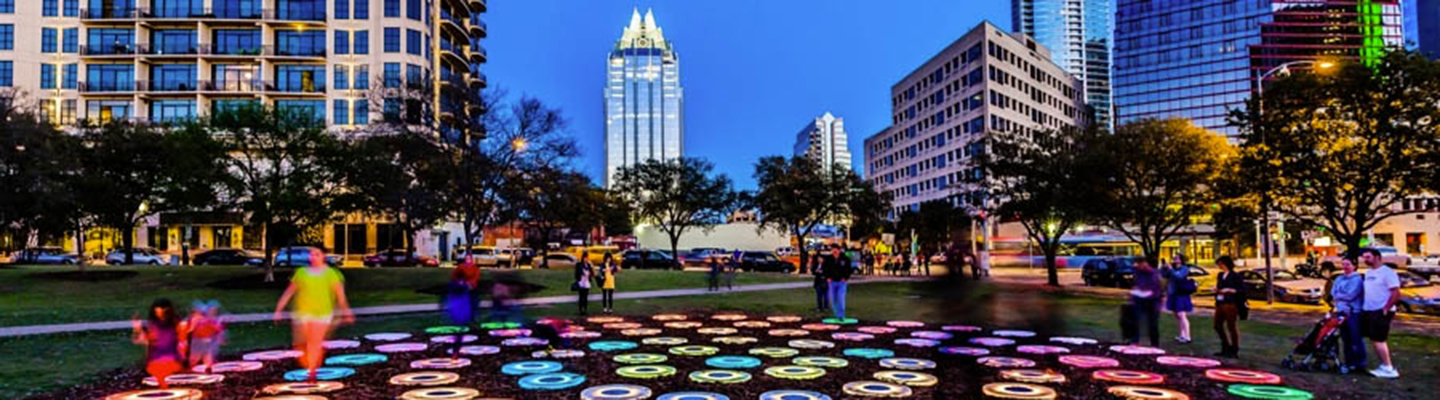 People in a park with a light up art display and downtown view in the back