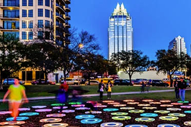 People in a park with a light up art display and downtown view in the back